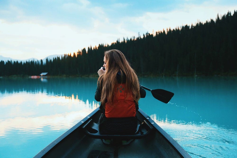 que ver en canada en 15 dias chica rubia de espaldas en una canoa remando en un lago con el agua transparente y montañas a su alrededor